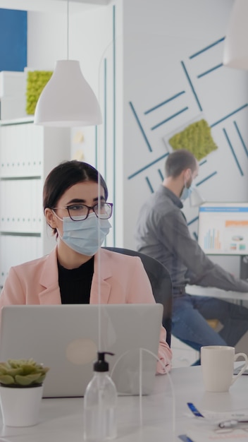 Arbeiter, die eine schützende Gesichtsmaske tragen, sprechen über das Tippen von Geschäftsprojekten auf dem PC im Büro während der globalen Pandemie des Coronavirus