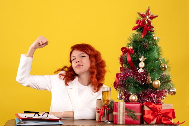 Arbeiterin, die hinter Tisch mit Weihnachtsbaum sitzt und auf Gelb präsentiert