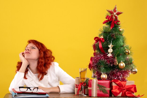 Arbeiterin, die hinter Tisch mit Weihnachtsbaum sitzt und auf Gelb präsentiert
