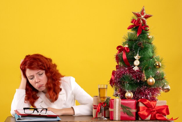 Arbeiterin, die hinter Tisch mit Weihnachtsbaum sitzt und auf Gelb präsentiert