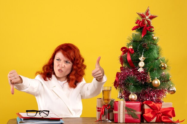 Arbeiterin, die hinter Tisch mit Weihnachtsgeschenken und Baum auf Gelb sitzt