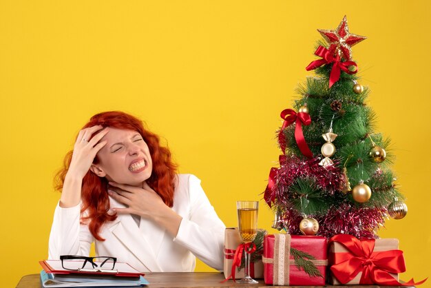 Arbeiterin, die hinter Tisch mit Weihnachtsgeschenken und Baum auf Gelb sitzt