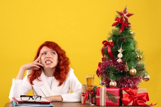 Arbeiterin, die hinter Tisch mit Weihnachtsgeschenken und Baum auf Gelb sitzt