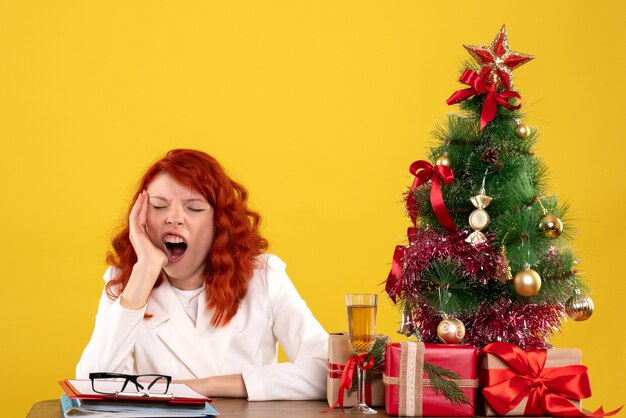 Arbeiterin, die hinter Tisch mit Weihnachtsgeschenken und Baum auf Gelb sitzt