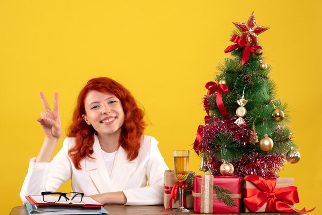 Arbeiterin, die hinter Tisch mit Weihnachtsgeschenken und Baum auf Gelb sitzt