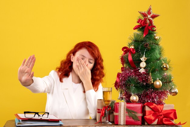 Arbeiterin, die hinter Tisch mit Weihnachtsgeschenken und Baum auf Gelb sitzt