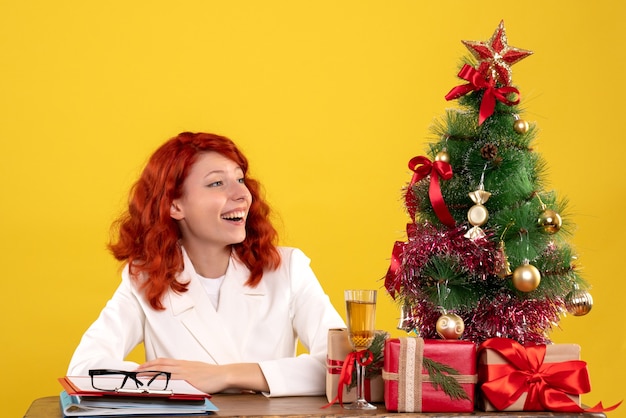 Arbeiterin, die hinter Tisch mit Weihnachtsgeschenken und Baum auf Gelb sitzt