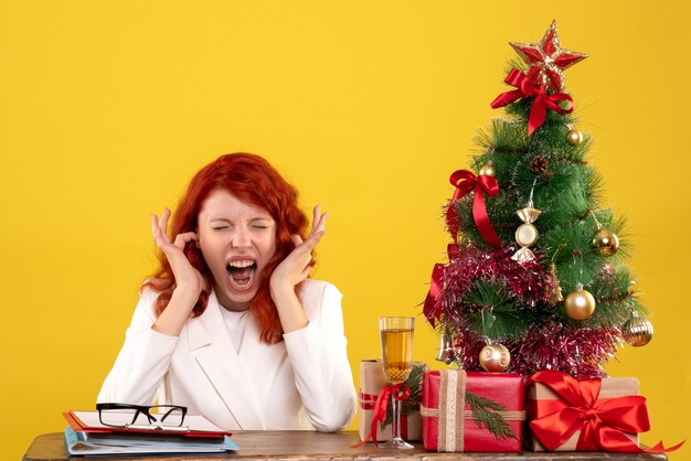 Arbeiterin, die hinter Tisch mit Weihnachtsgeschenken und Baum auf Gelb sitzt