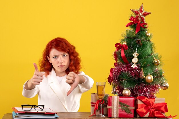 Arbeiterin, die hinter Tisch mit Weihnachtsgeschenken und Baum auf Gelb sitzt