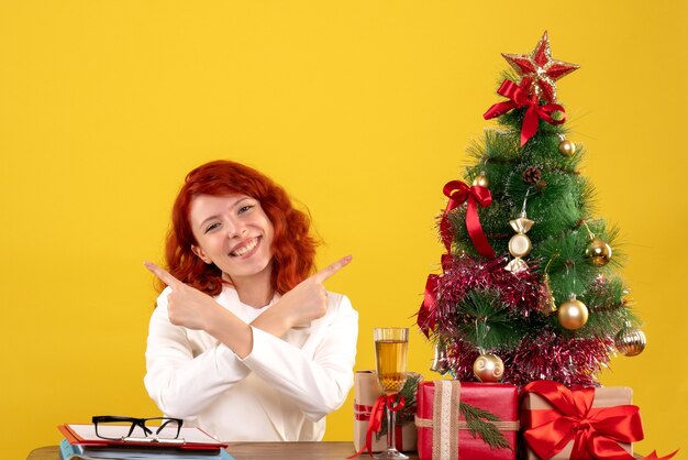 Arbeiterin, die hinter Tisch mit Weihnachtsgeschenken und Baum auf gelbem Schreibtisch sitzt