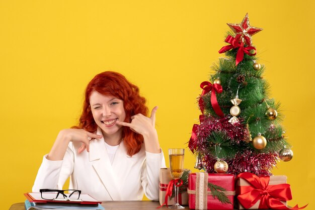 Arbeiterin, die hinter Tisch mit Weihnachtsgeschenken und Baum sitzt, der auf Gelb lächelt