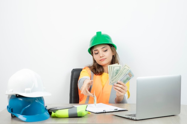 Arbeiterin in orangefarbener weste und grünem helm sitzt am schreibtisch. foto in hoher qualität