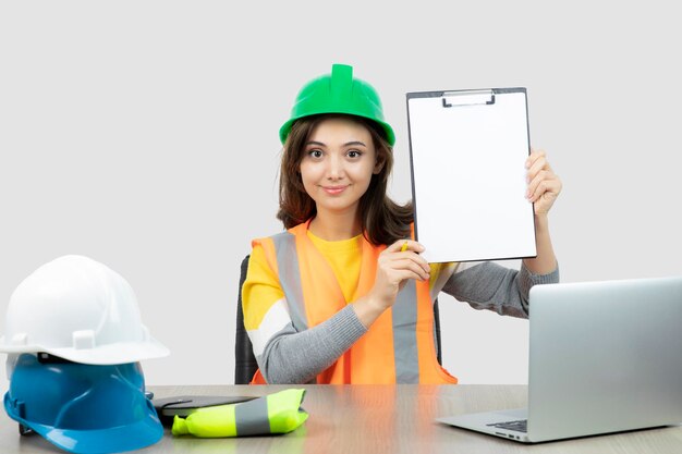 Arbeiterin in Uniform sitzt mit Laptop und Klemmbrett am Schreibtisch. Foto in hoher Qualität