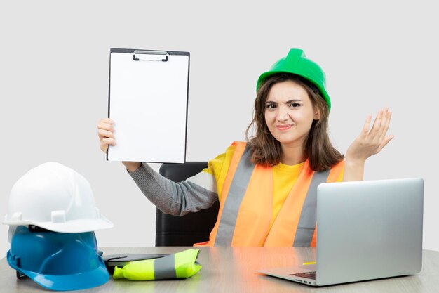 Arbeiterin in Uniform sitzt mit Laptop und Klemmbrett am Schreibtisch. Foto in hoher Qualität