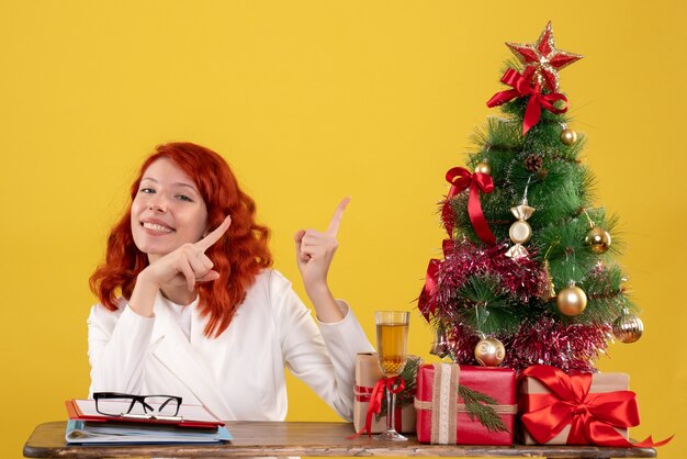 Arbeiterin sitzt hinter ihrem Tisch mit Weihnachtsbaum und präsentiert auf gelb
