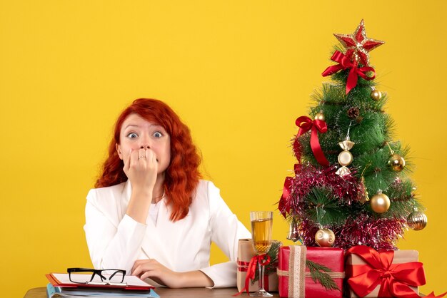 Arbeiterin sitzt hinter Tisch mit Weihnachtsgeschenken und Baum nervös auf gelb