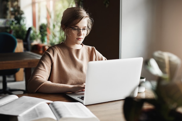 Arbeits-, Lifestyle- und Geschäftskonzept. Gut aussehende fokussierte europäische Frau in trendigen Gläsern, die im Café nahe Laptop sitzen, am Notizbuch arbeiten, mit Büchern umgeben, Notizen machen.