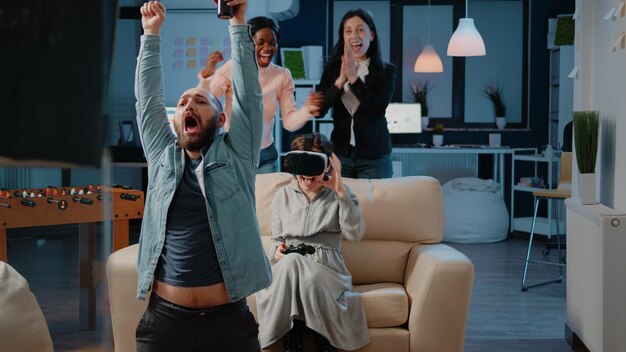 Arbeitskollegen, die nach der Arbeit im Büro Videospiele mit Controller auf der Konsole spielen. Frau gewinnt Spiel mit VR-Brille und Joystick im Fernsehen und genießt lustige Aktivitäten mit Getränken.