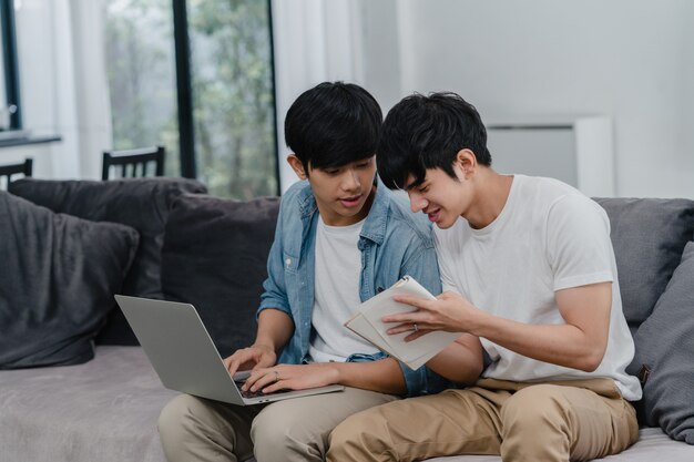 Arbeitslaptop der jungen asiatischen homosexuellen Paare am modernen Haus. Die glücklichen Männer Asiens LGBTQ + entspannen sich den Spaß, der Computer verwendet und zusammen ihre Finanzen im Internet analysiert, während Lügensofa im Wohnzimmer am Haus.