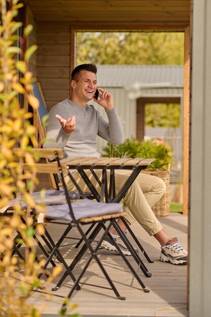 Arbeitsleben. Fröhlicher Mann mittleren Alters in bequemer Freizeitkleidung, der auf dem Smartphone gestikuliert, während er an einem sonnigen Tag auf der offenen Terrasse des Landhauses sitzt