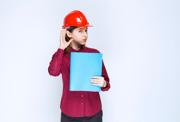 Architektin im roten Helm mit blauem Ordner und posiert vor der Kamera.