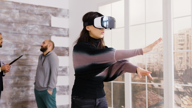 Architektin mit Virtual-Reality-Brille in leerer Wohnung und Immobilienmakler im Gespräch mit dem Kunden im Hintergrund.