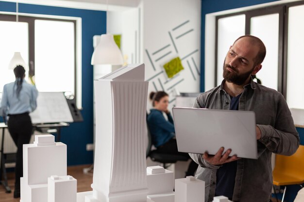 Architekturinspektor mit Laptop-Neigungskopf und Blick auf die Maquette des Wolkenkratzers in einem Wohnprojekt. Architekt, der über gestalterische Verbesserungen am maßstabsgetreuen Modell der Stadtplanung nachdenkt.