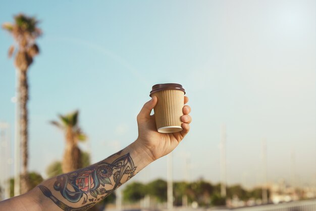 Arm und Hand des tätowierten Mannes mit einer beige Einwegkaffeetasse aus Wellpappe gegen klaren blauen Himmel und Palmen
