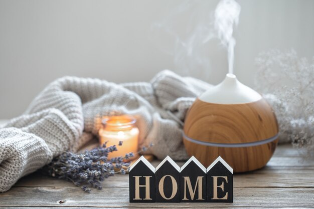 Aroma Stillleben mit einem modernen Aromaöldiffusor auf einer Holzoberfläche mit einem gestrickten Element, gemütlichen Details und dem dekorativen Wort Zuhause.