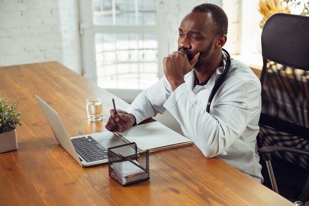 Arzt berät Patienten, hört gestresster afroamerikanischer Arzt während seiner Arbeit mit Patienten zu und erklärt Rezepte für Medikamente. Tägliche harte Arbeit für Gesundheit und Leben retten während der Epidemie.