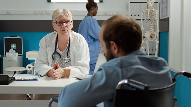 Kostenloses Foto arzt berät rollstuhlfahrer in einem behindertenfreundlichen büro, um chronische gesundheitsprobleme zu heilen. mann mit körperlicher beeinträchtigung, der an einem kontrolltermin teilnimmt, rat und unterstützung erhält.