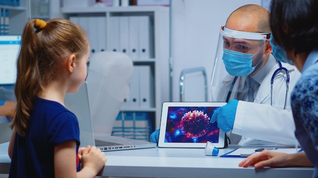 Arzt, der die Entwicklung des Virus mit Schutzmaske mit Tablet erklärt. Kinderarztspezialist mit Visier und Handschuhen, der Gesundheitsdienste, Beratungen und Behandlungen während des Coronavirus anbietet.