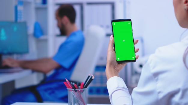 Arzt, der Smartphone mit Modell im Krankenhausschrank hält, während Krankenpfleger am Computer arbeitet. Gesundheitsspezialist im Krankenhausschrank mit Smartphone mit Modell.