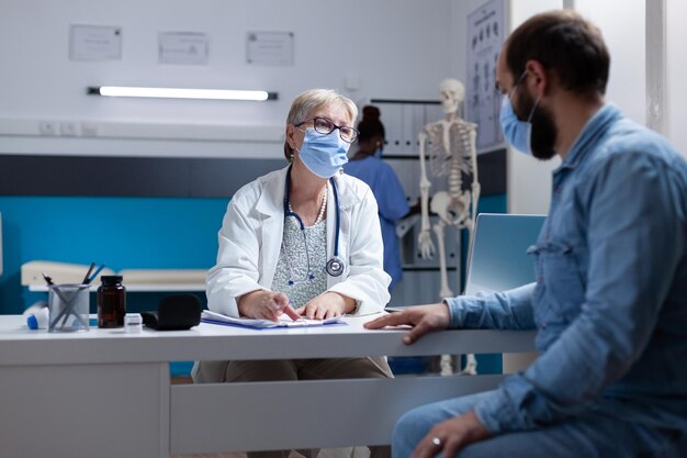 Arzt, der während der Covid-19-Pandemie im Kabinett Beratung mit einem kranken Mann macht. Ärztin, die den Patienten untersucht und bei der Gesundheitsbehandlung hilft, wobei sie beim Kontrollbesuch eine Gesichtsmaske trägt.