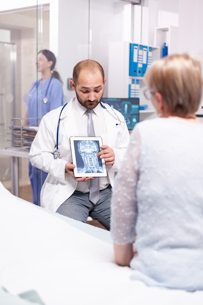 Arzt erklärt einer älteren Frau auf einem Tablet-PC im Untersuchungsraum des Krankenhauses eine Gehirnverletzung?