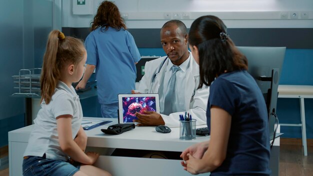 Arzt erklärt Erwachsenen und kleinen Kindern im medizinischen Kabinett die Coronavirus-Illustration auf dem Tablet-Display. Mann, der bei der Untersuchung über Virenverbreitungsanimation auf dem Gerät spricht.