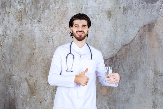 Arzt mit einem Stethoskop, der ein Glas reines Wasser hält und ein positives Handzeichen zeigt