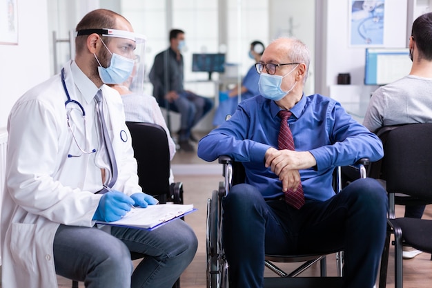 Arzt mit Gesichtsmaske und Stethoskop, der einen behinderten älteren Mann im Wartebereich des Krankenhauses berät