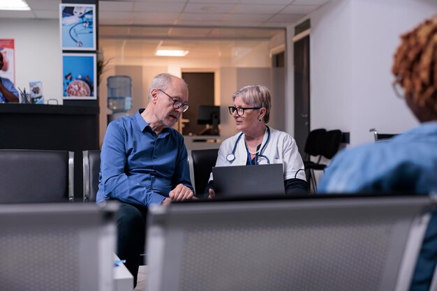 Arzt mit Laptop bei Untersuchungsbesuch mit altem Mann, der in der Lobby des Wartezimmers sitzt. Hausarzt und Patient sprechen über Krankheitsdiagnose und Gesundheitsversorgung und konsultieren Erwachsene im Gesundheitszentrum.