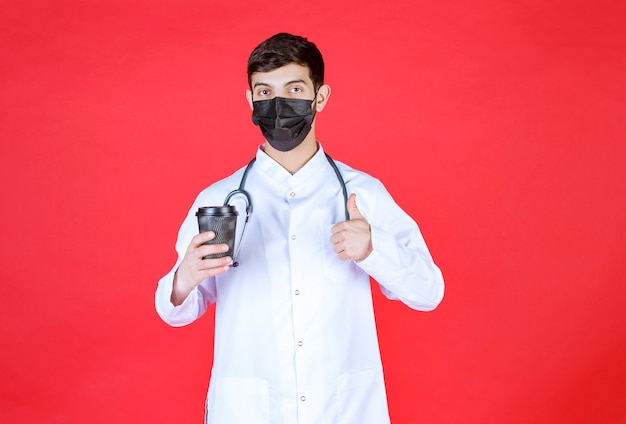 Arzt mit stethoskop in schwarzer maske, der eine schwarze kaffeetasse zum mitnehmen hält und den geschmack genießt.