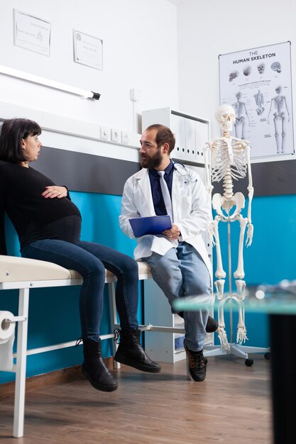 Arzt mit Zwischenablage, der schwangere Frau beim Prüfungstermin im Büro berät. Werdende Patientin bespricht mit dem Arzt über Geburt und Schwangerschaft und wird beim Kontrollbesuch unterstützt.