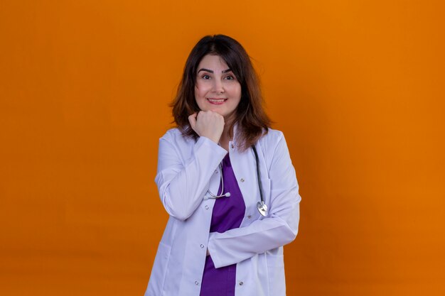Arzt mittleren Alters, der weißen Kittel trägt und mit Stethoskop fröhlich mit Hand nahe Kinn über isolierte orange Wand lächelt
