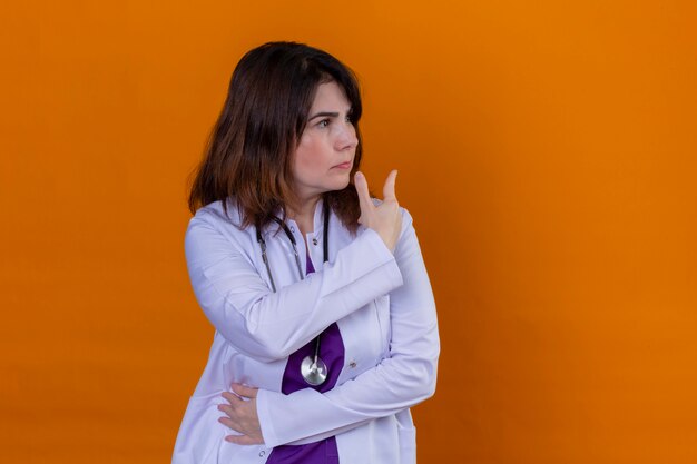 Arzt mittleren Alters in weißem Kittel und mit Stethoskop, das mit Hand und Finger nach hinten zeigt, missfallen über der isolierten orangefarbenen Wand