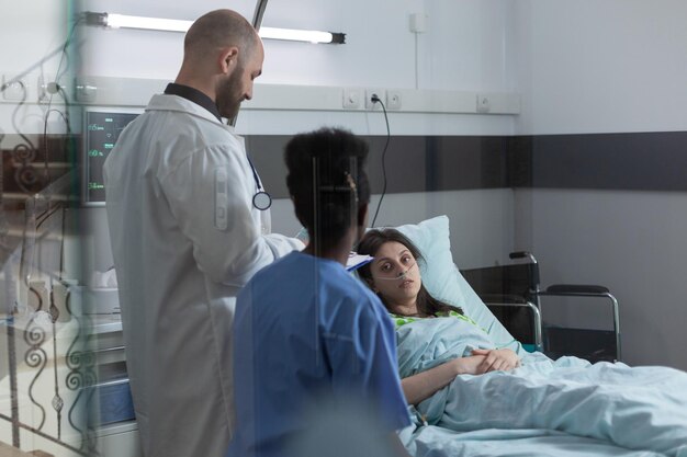 Arzt und Krankenschwester beraten sich in der Privatabteilung, um die Frau nach einem chirurgischen Eingriff im Krankenhausbett zu erholen. Patient mit Nasenkanüle, der medizinisches Fachpersonal anschaut.