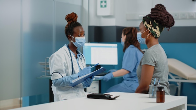 Arzt und patient mit gesichtsmasken sprechen bei der jährlichen untersuchung zur untersuchung über krankheiten. Sanitäter berät junge Frau im Kabinett, während sie Schutz vor Coronavirus hat