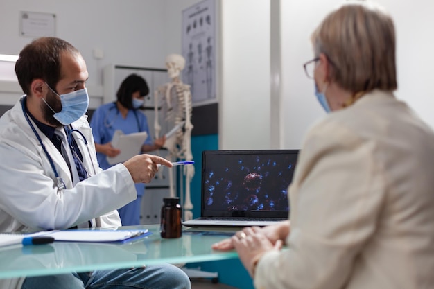 Arzt zeigt auf Coronavirus-Präsentation, um dem reifen Patienten beim jährlichen Kontrollbesuch Bakterien zu zeigen. Ältere Frau und Mediziner, die Virusillustration auf Laptop im Schrank analysieren.