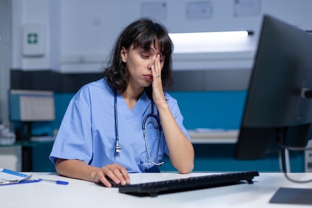 Arzthelferin, die einschläft, während sie Computer und Tastatur für das Gesundheitswesen verwendet. Frauenkrankenschwester, die den Monitor auf dem Schreibtisch verwendet, sich erschöpft fühlt und spät in der Nacht arbeitet. Müde Praktiker