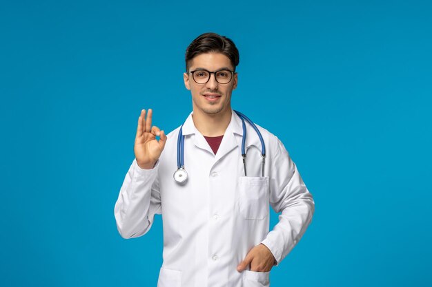 Arzttag hübscher brünetter süßer kerl im medizinischen kleid, der ok-zeichen mit stethoskop zeigt