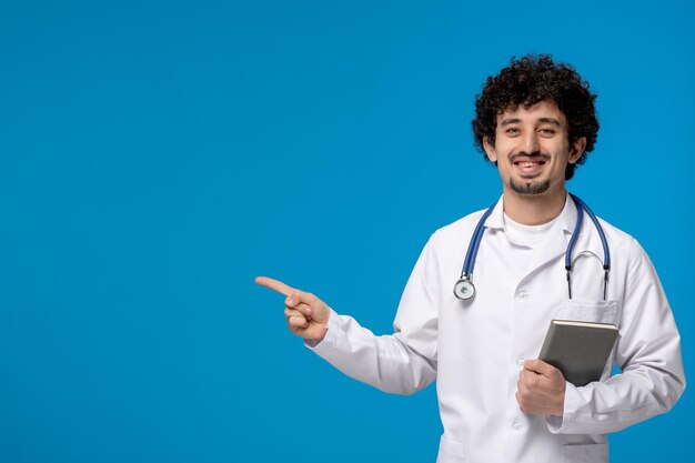 Arzttag, lockiger, brünetter, süßer Kerl in medizinischer Uniform, der lächelt und ein Buch hält