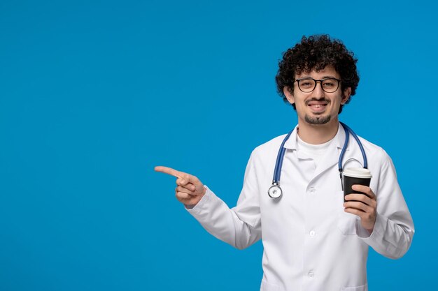 Arzttag lockiger, gutaussehender, süßer Kerl in medizinischer Uniform, der eine Brille trägt und eine Kaffeetasse in der Hand hält